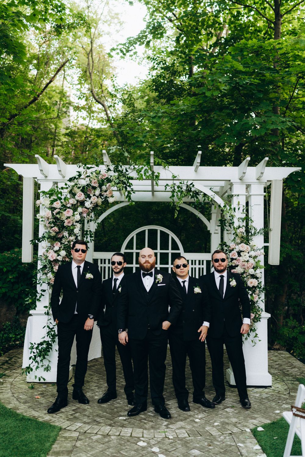 A group in black suits and sunglasses stand under a white floral wedding arch in a garden setting.