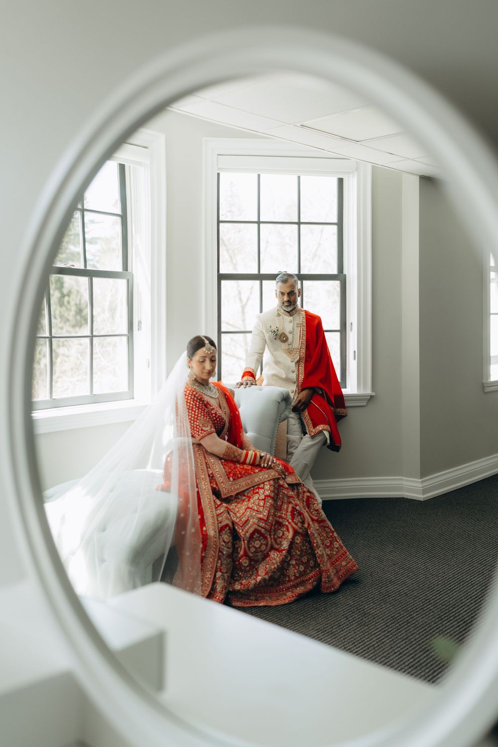 A mirror reflection shows a couple in South Asian wedding attire posing by a window in an elegant room.