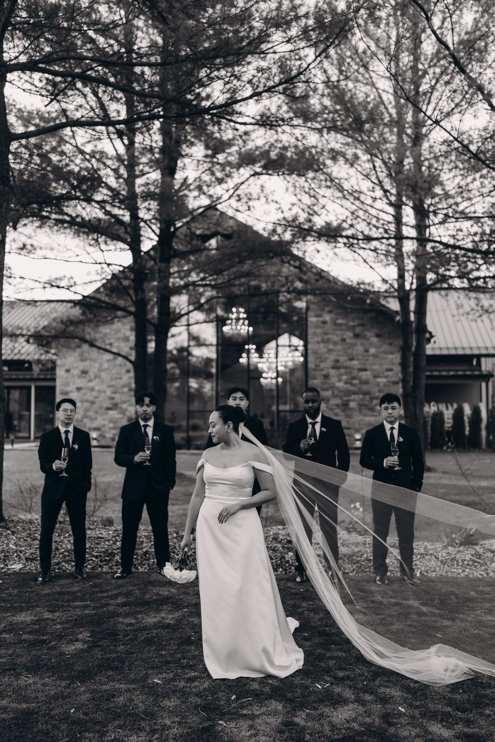 Wedding party poses in black and white photo with dramatic flowing veil against brick building backdrop.
