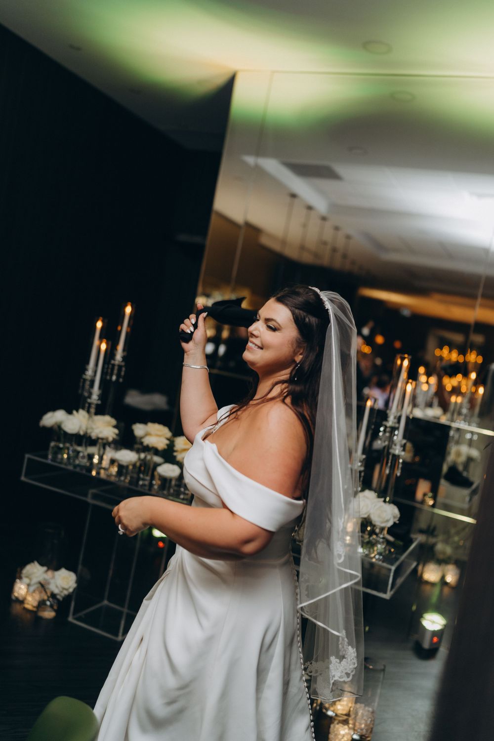 Bride in off-shoulder white gown and veil stands among candlelit decor during elegant evening reception.