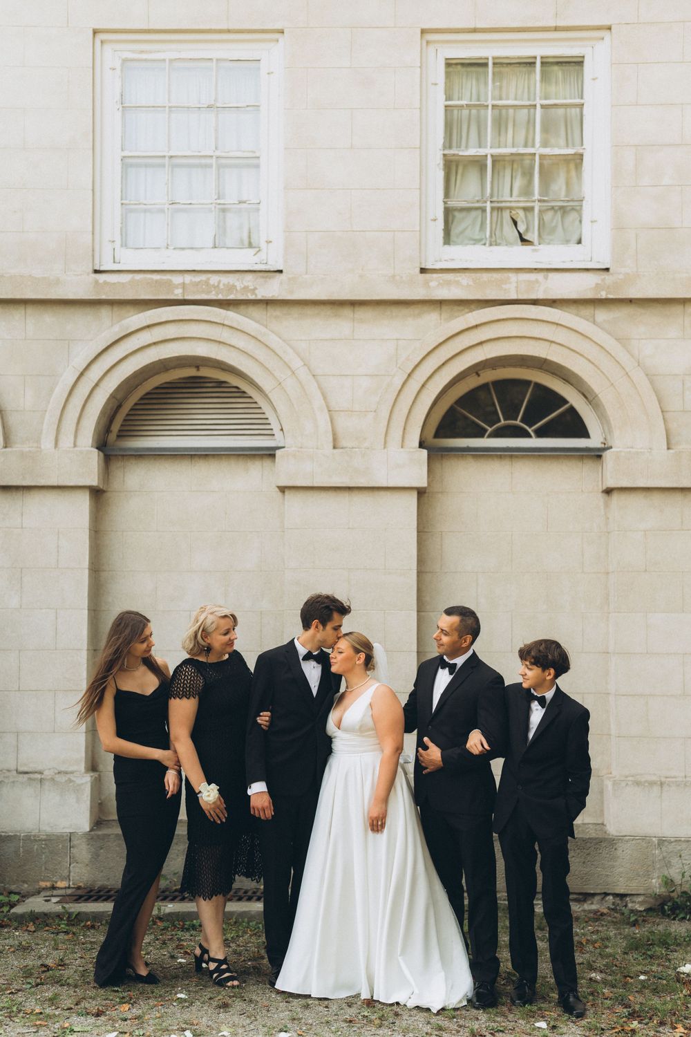 Wedding party in formal attire stands together against classic architectural building with arched windows and doorways.