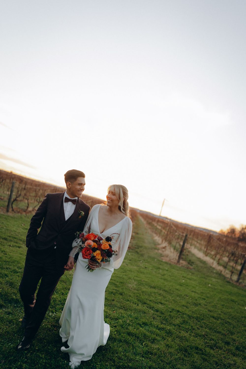 A couple shares a romantic moment in their wedding attire at sunset in a vineyard with autumn-colored flowers.