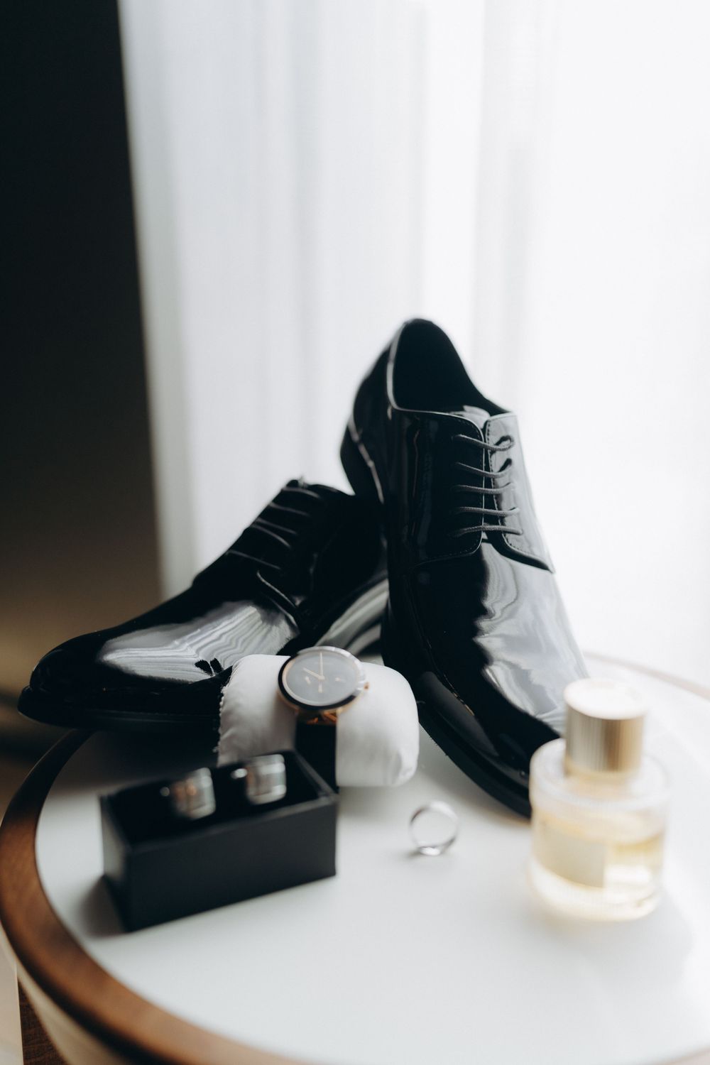 Elegant black leather dress shoes styled with cufflinks and perfume bottle on white surface in moody lighting.