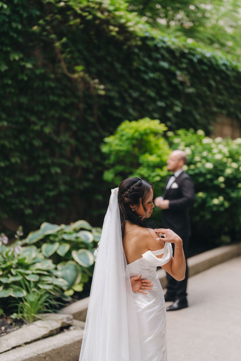 A bride in a white strapless wedding dress and long veil stands in a garden pathway surrounded by lush greenery.