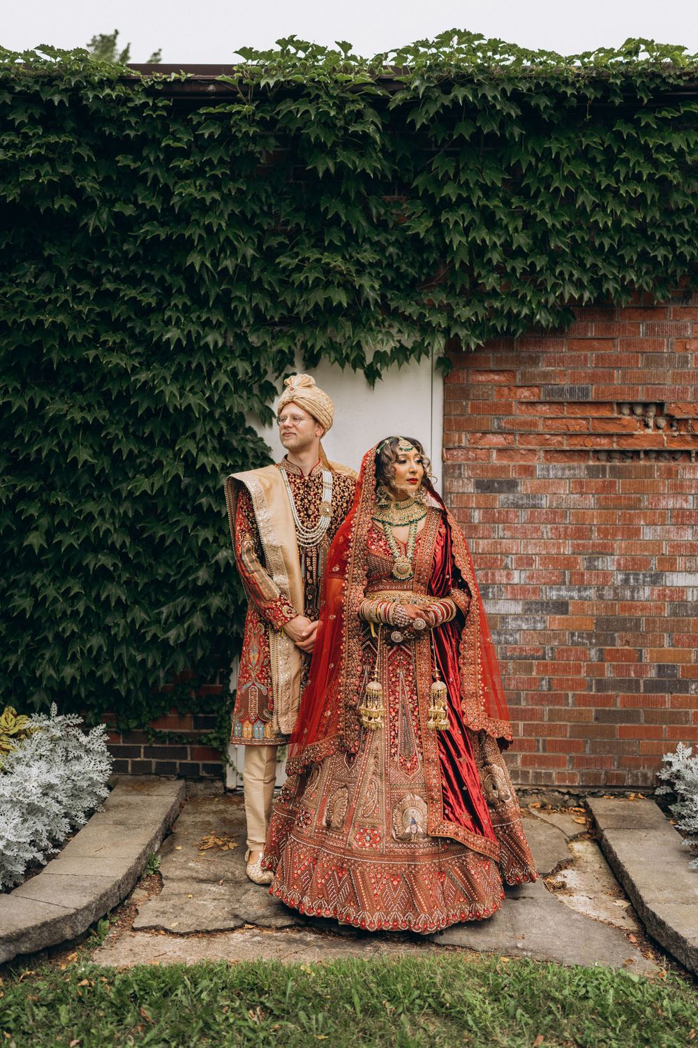 Traditional Indian wedding couple in ornate red and gold attire stand against brick wall with green ivy backdrop.