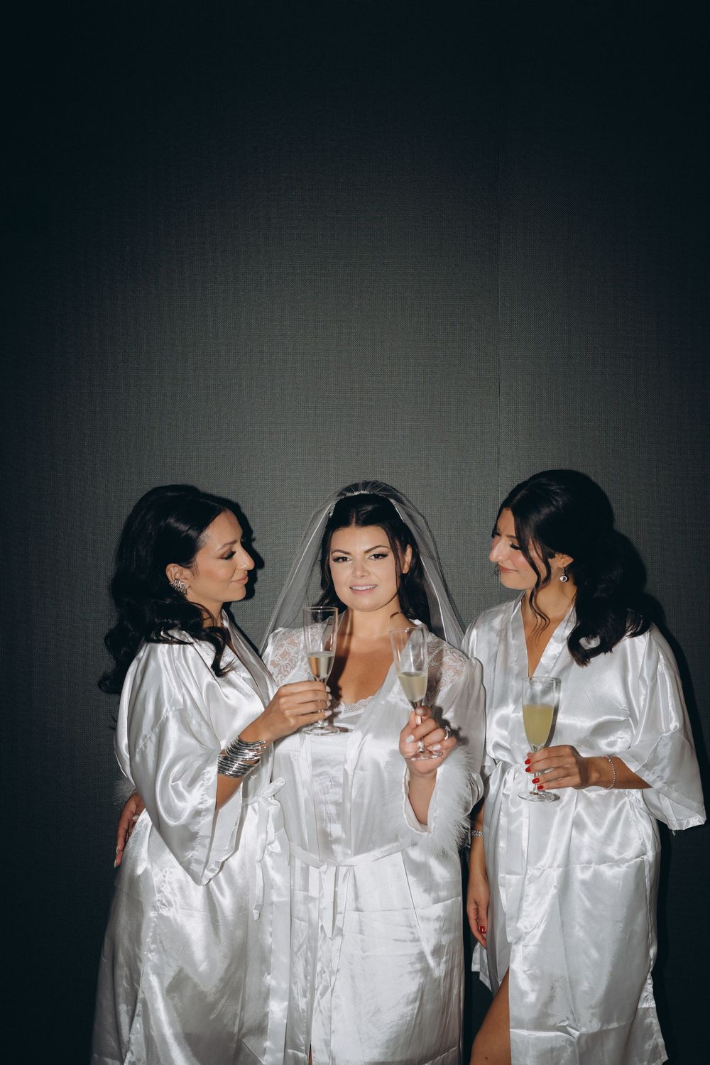 Three women in matching white silk robes stand closely together with champagne glasses during a celebration.