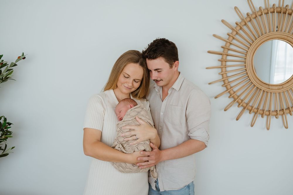 A couple tenderly holds their newborn baby, standing against a soft blue wall with a rattan mirror.