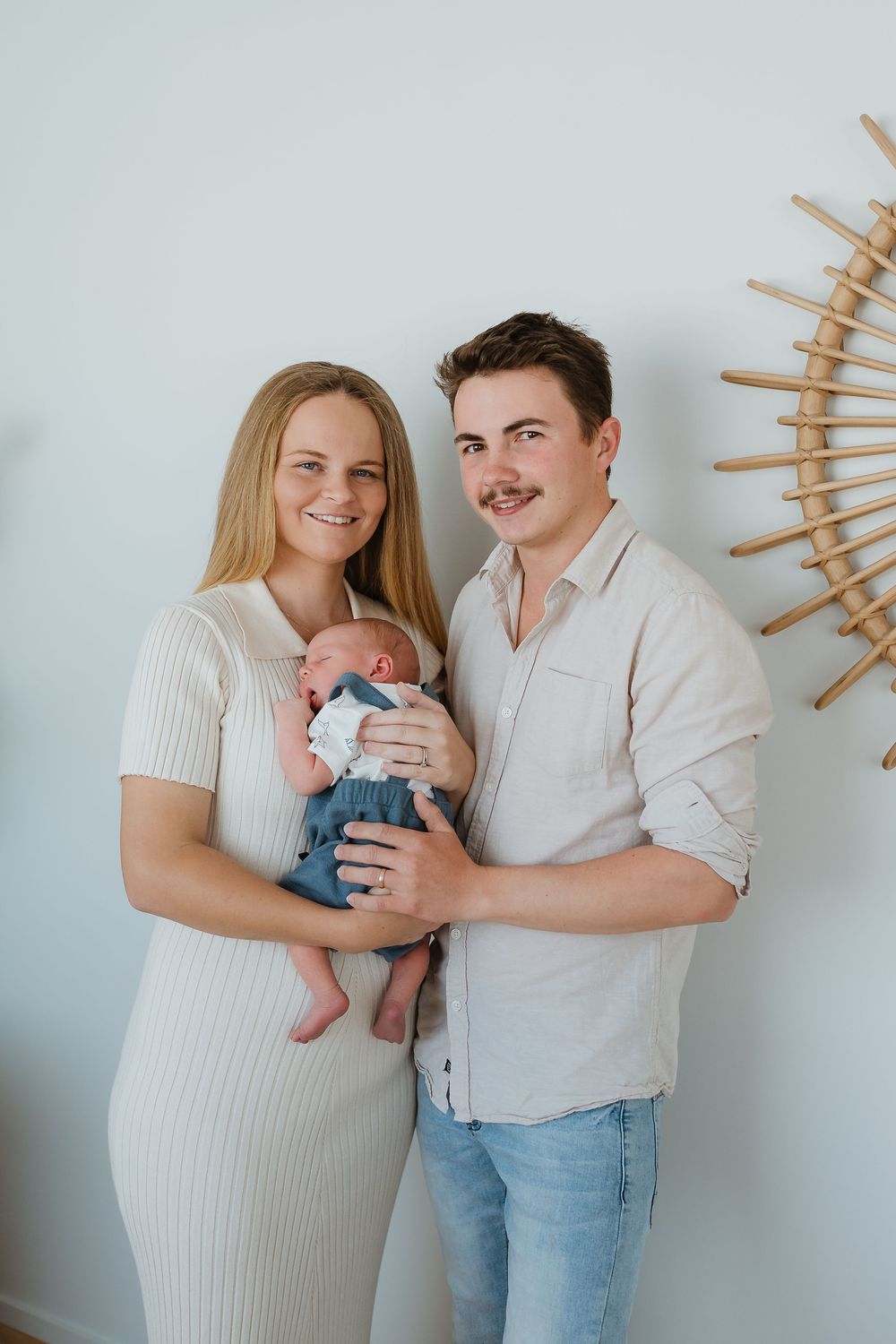 Young couple smiling and holding their newborn baby, dressed in casual attire, standing against a light wall.