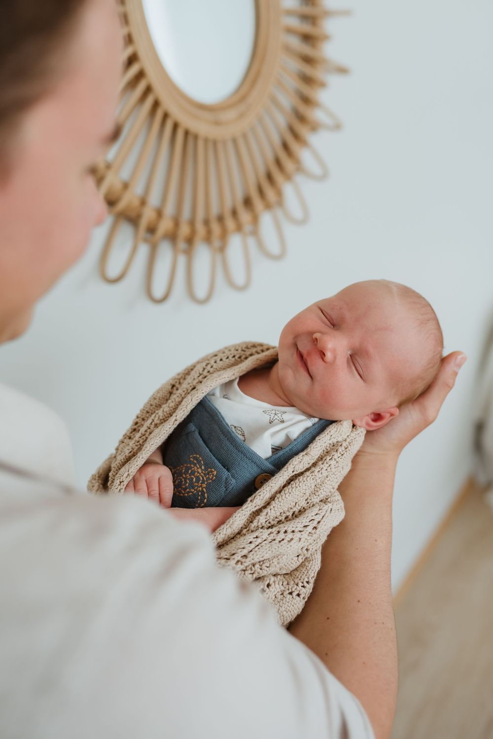Mother cradling sleeping newborn baby wrapped in a beige knit blanket and blue outfit indoors.