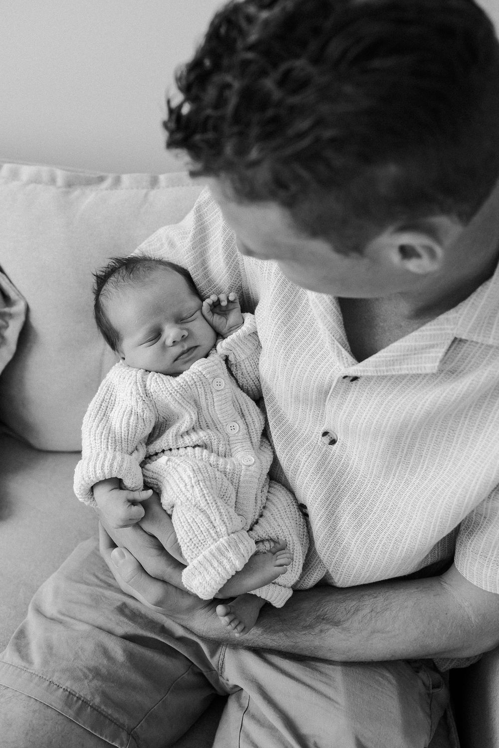 Black and white photo of a father cradling his sleeping newborn baby while sitting on a couch.