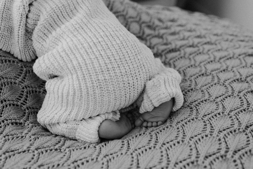 Black and white photo of a newborn baby's tiny feet peeking out from a cozy knit blanket and sweater.
