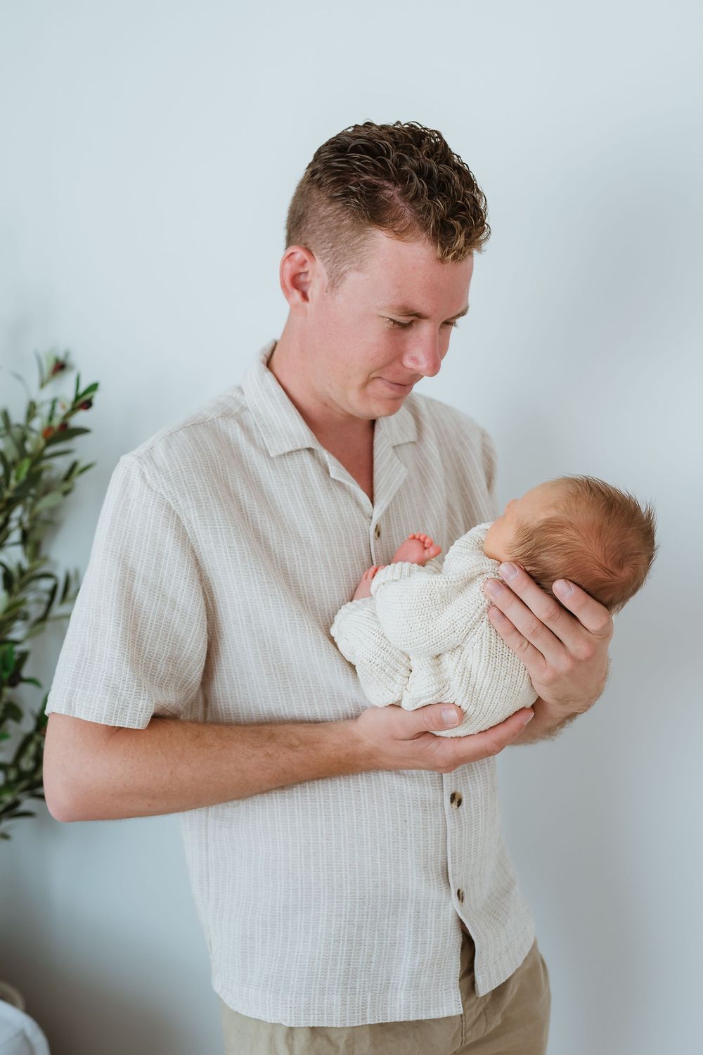 A father tenderly holds his newborn baby in both arms, gazing down lovingly while standing against a white wall.