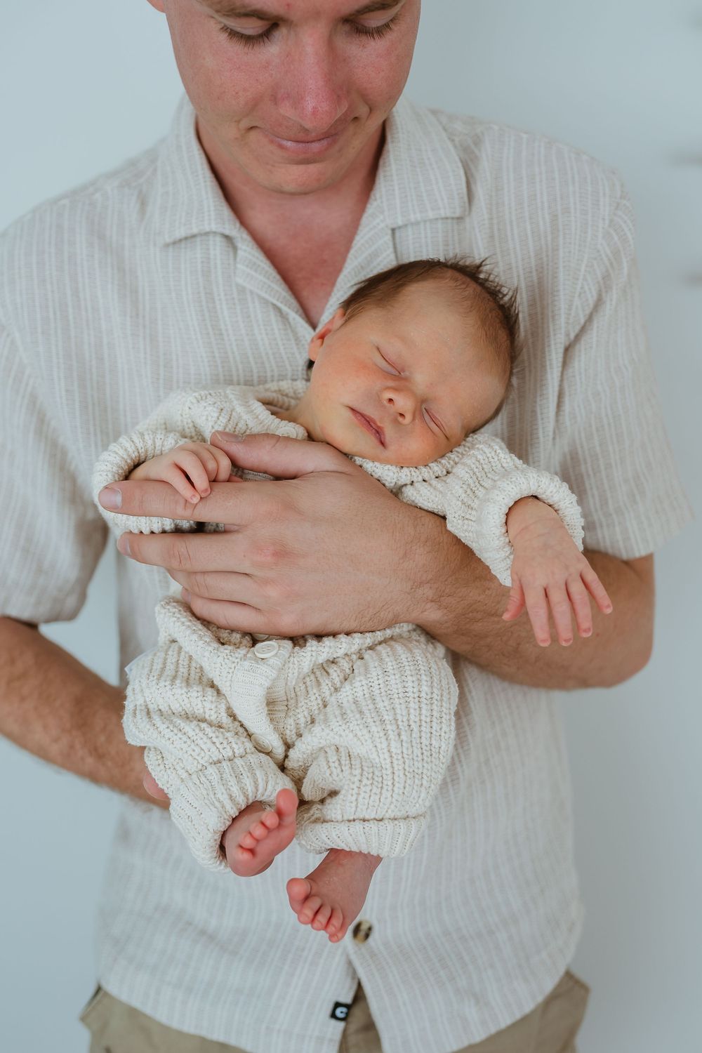 Father in beige shirt cradling sleeping newborn baby dressed in cream knit outfit against white background.
