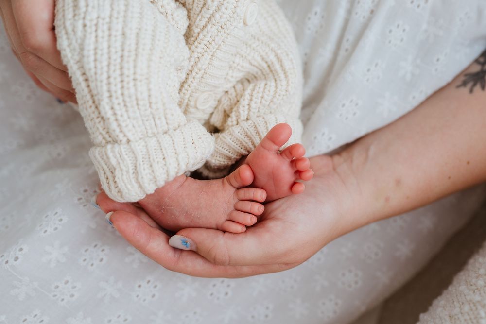 A parent gently cradles a newborn baby's tiny feet in their hands, wearing a cozy cream knit outfit.