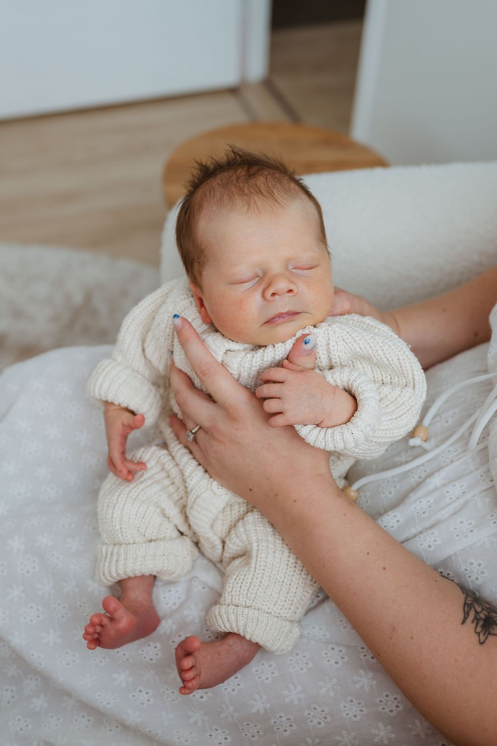 A newborn baby in a cream knit outfit sleeps peacefully while being gently held by a parent's hands.