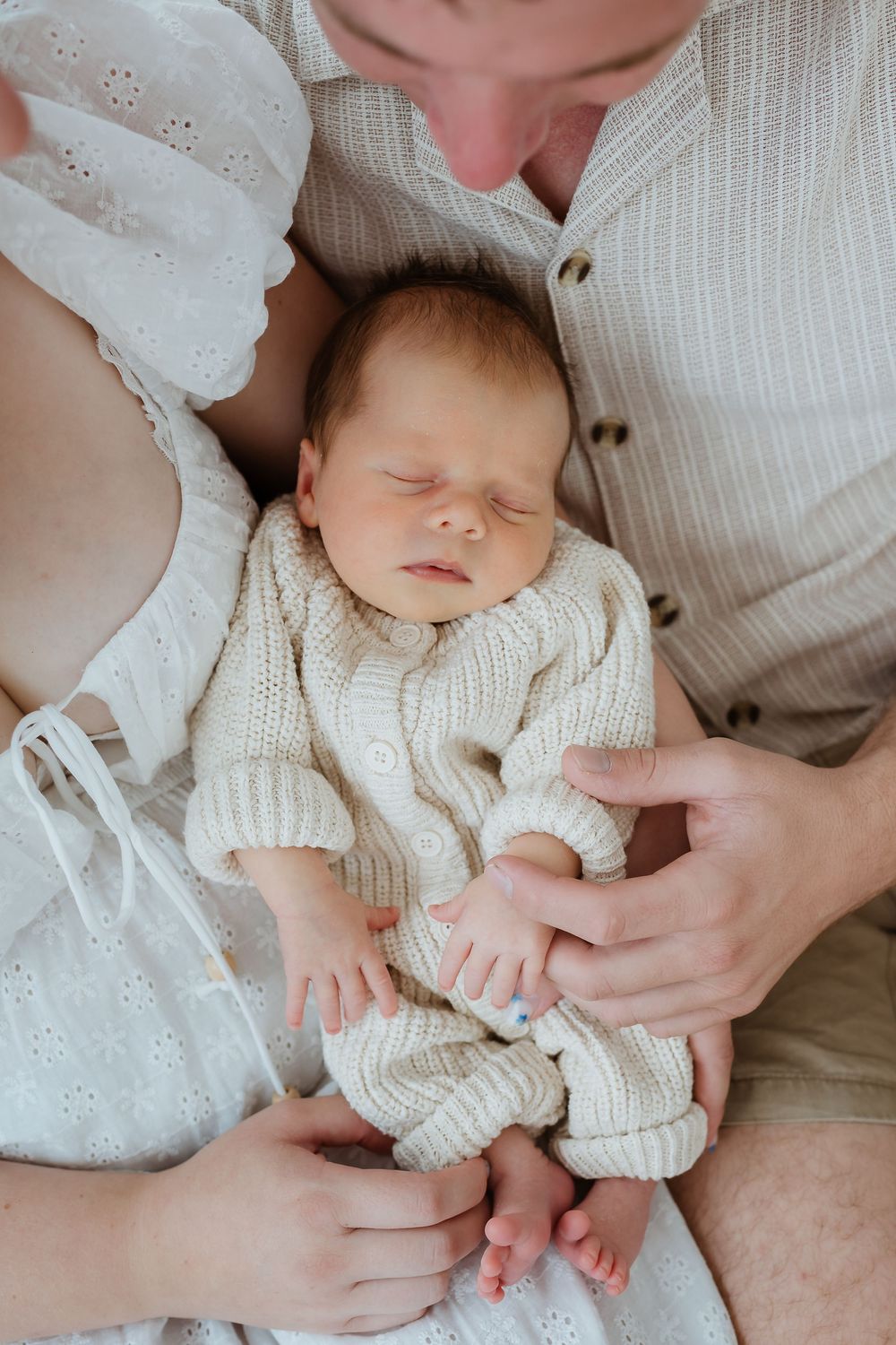 Sleeping newborn baby in cream knit onesie cradled in parents arms, peaceful family portrait.