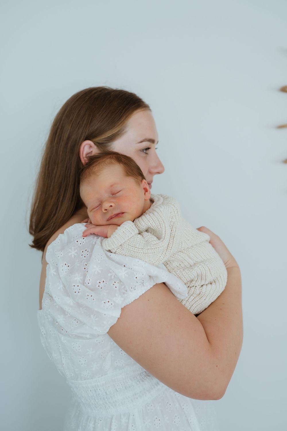 A young mother in a white dress gently holds her sleeping newborn baby against her shoulder.