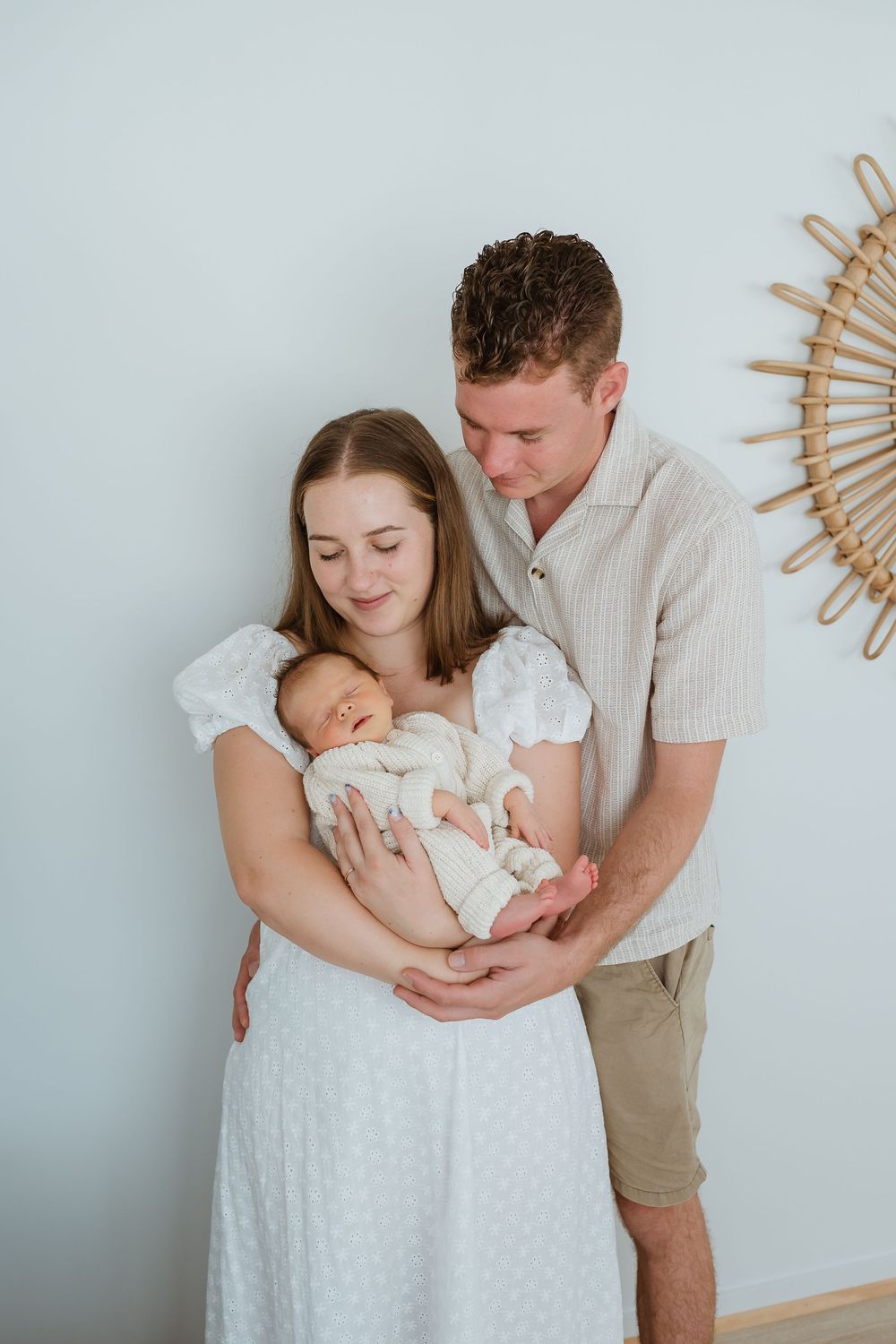 Young couple in white and beige outfits tenderly holding their sleeping newborn baby against a soft blue wall.
