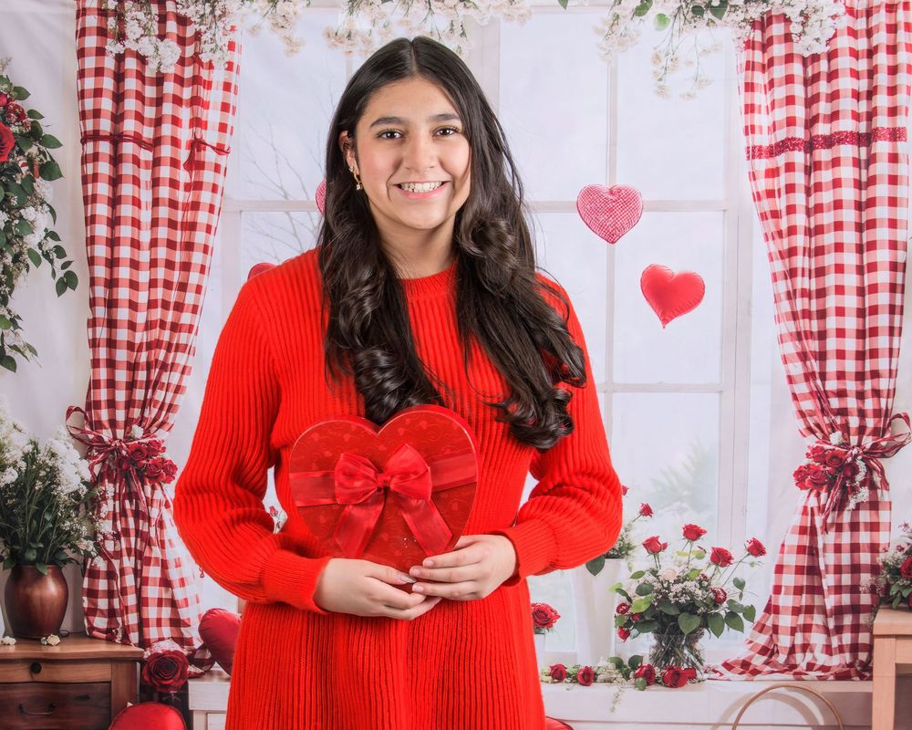 A person in a bright red sweater poses against a backdrop with red gingham curtains and heart decorations.