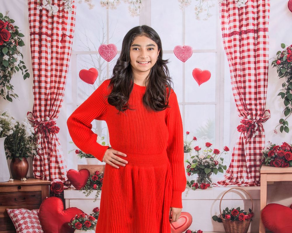 A person in a bright red sweater poses against a white wall with red heart decorations and checkered curtains.