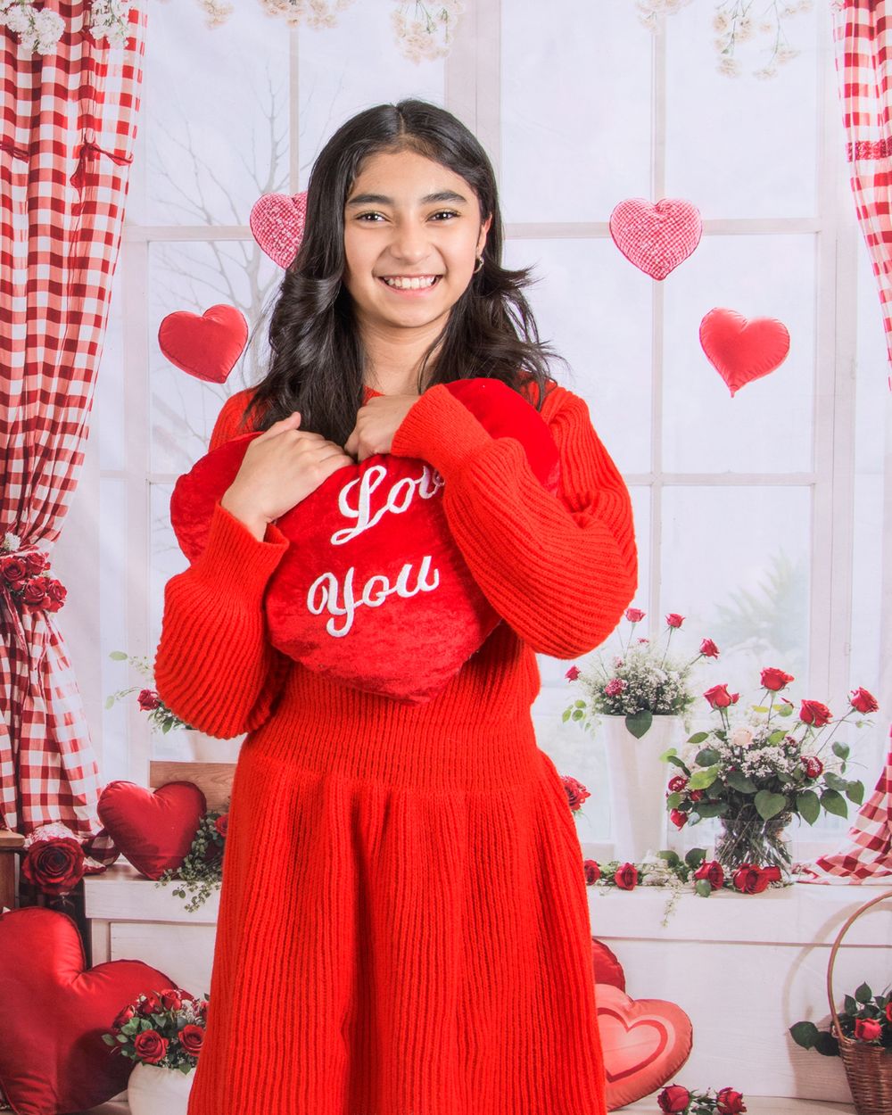 A person in a cozy red sweater poses in a Valentine's Day themed setting with checkered curtains and heart decorations.