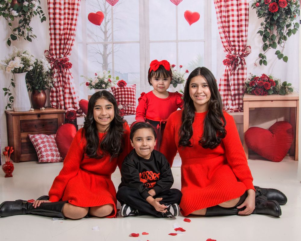 A group in matching red sweaters poses together in a festive holiday setting with gingham curtains and heart decorations.