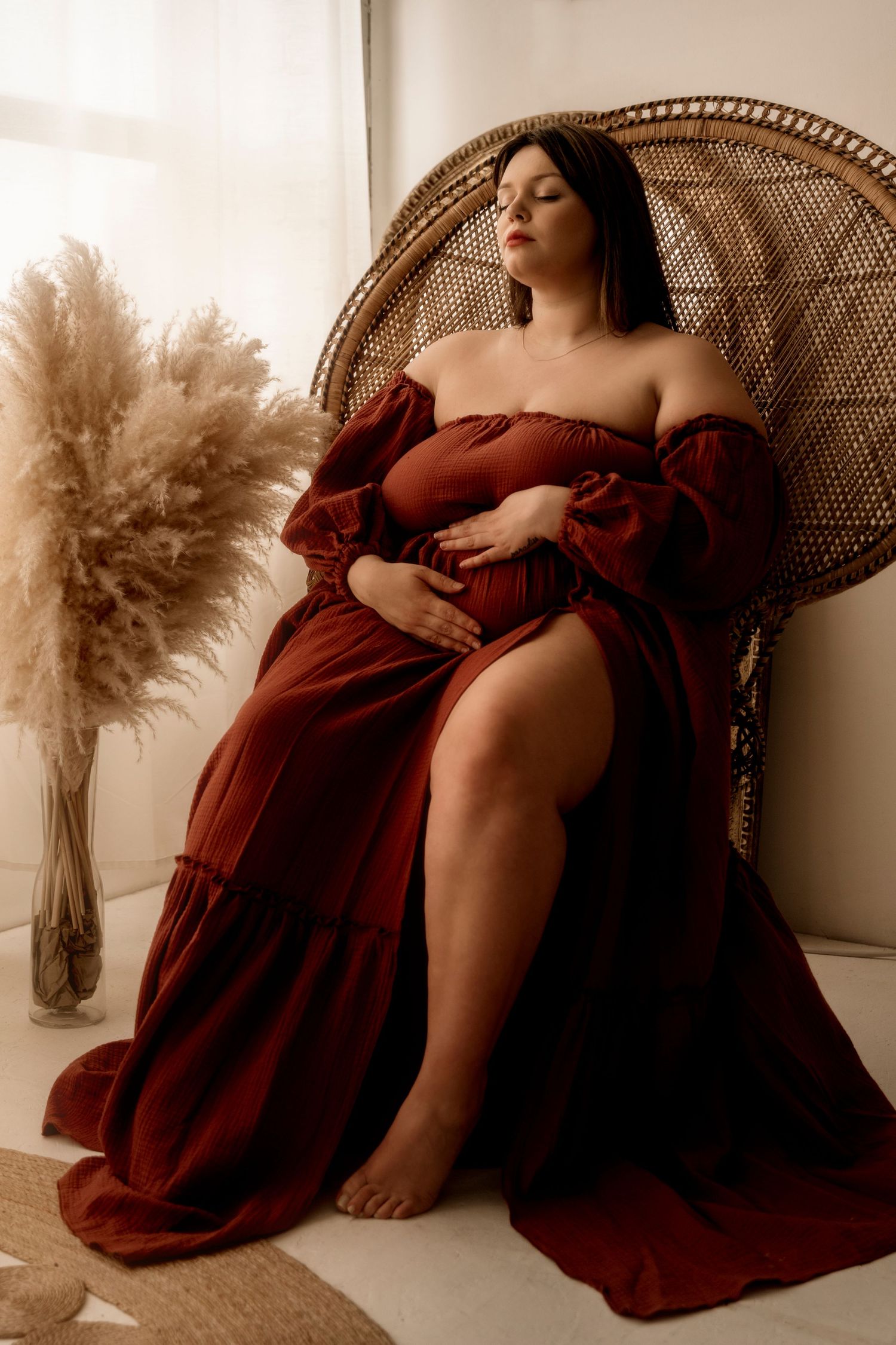 A feminine silhouette in a burgundy off-shoulder dress poses elegantly in a rattan peacock chair beside pampas grass decor.