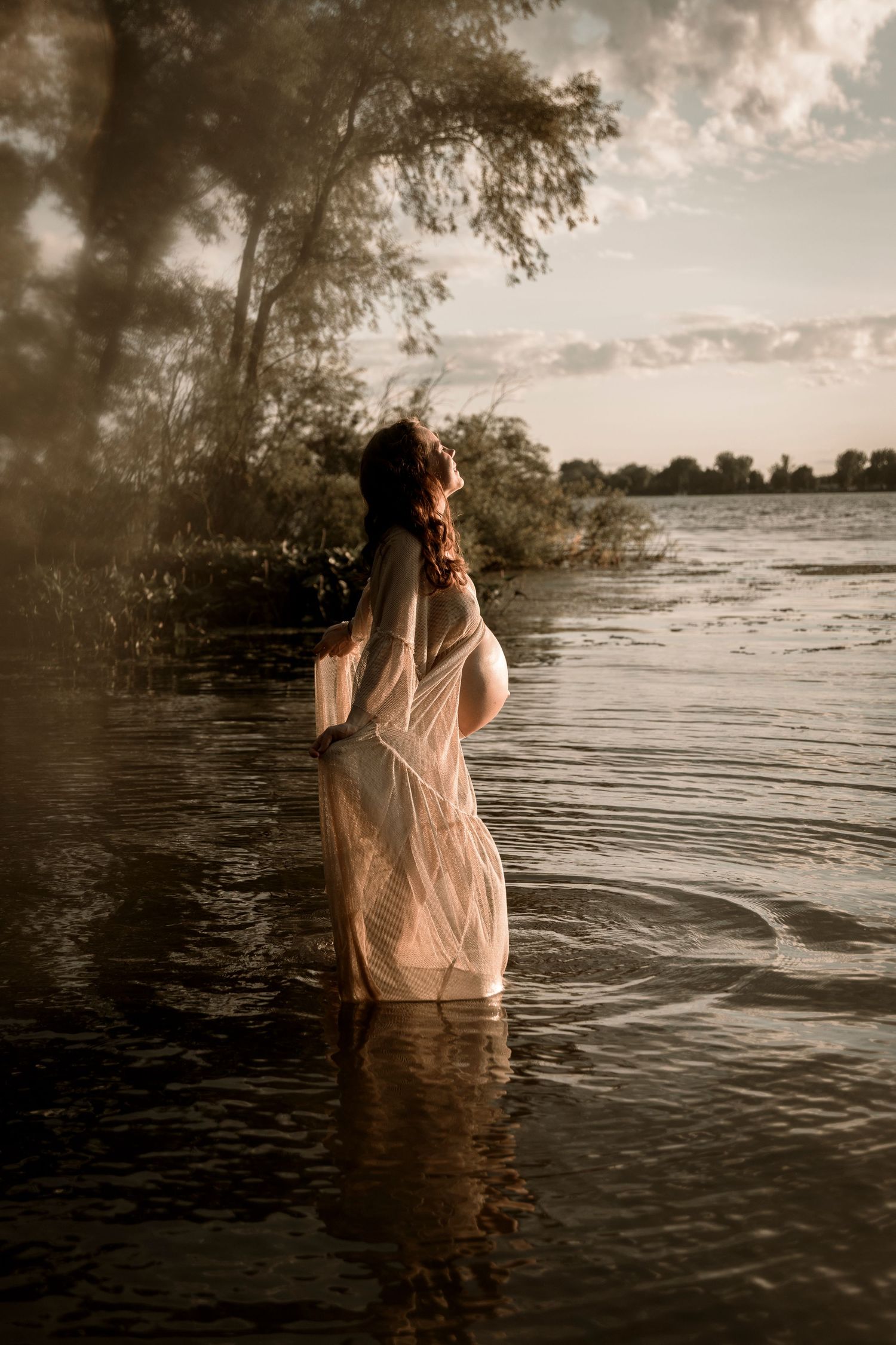 A dreamy figure in a flowy dress stands in rippling water at sunset, creating an ethereal silhouette against a cloudy sky.