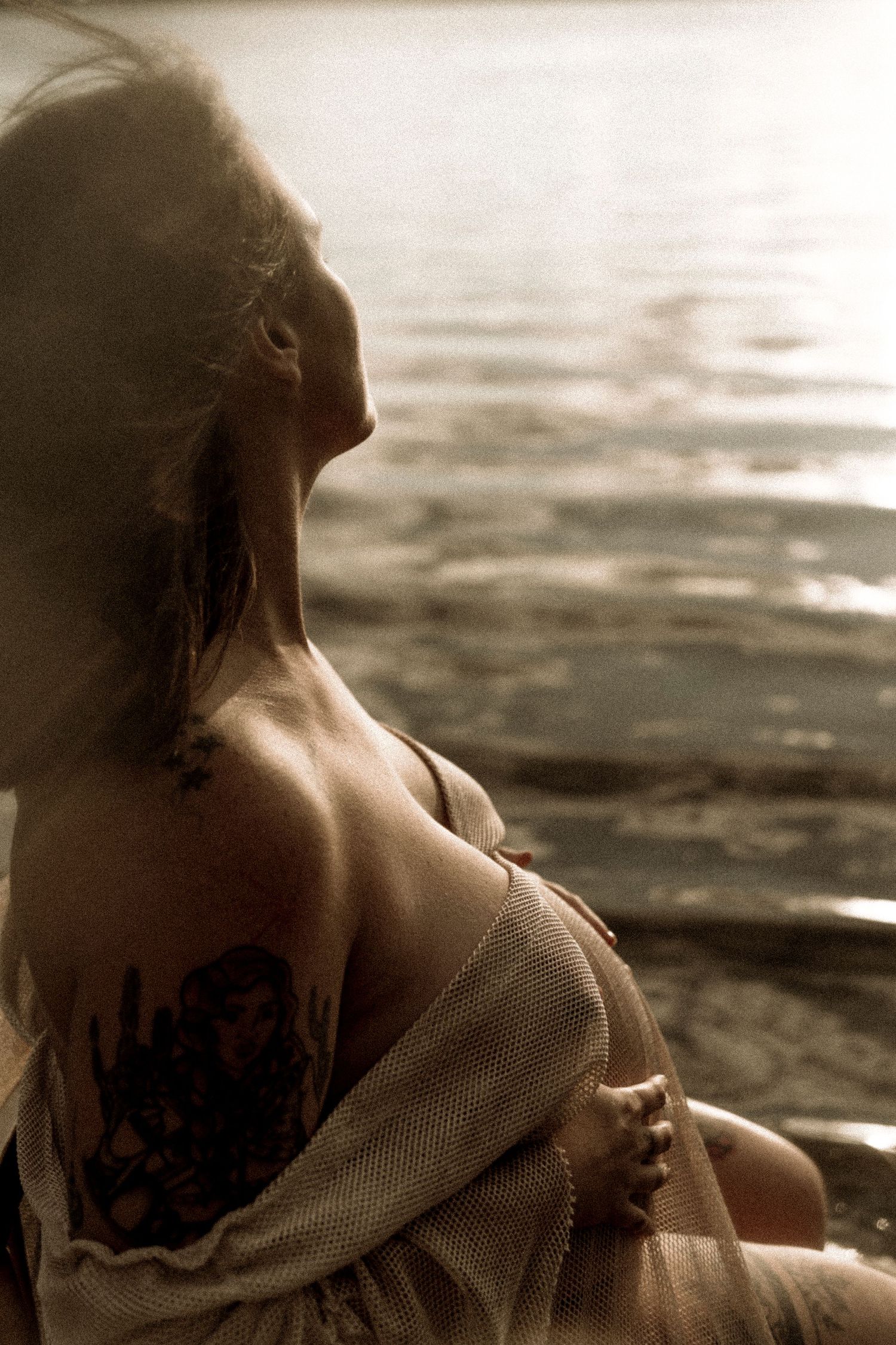Silhouette photo of a person with visible shoulder tattoo sitting by water at sunset in dreamy sepia tones.