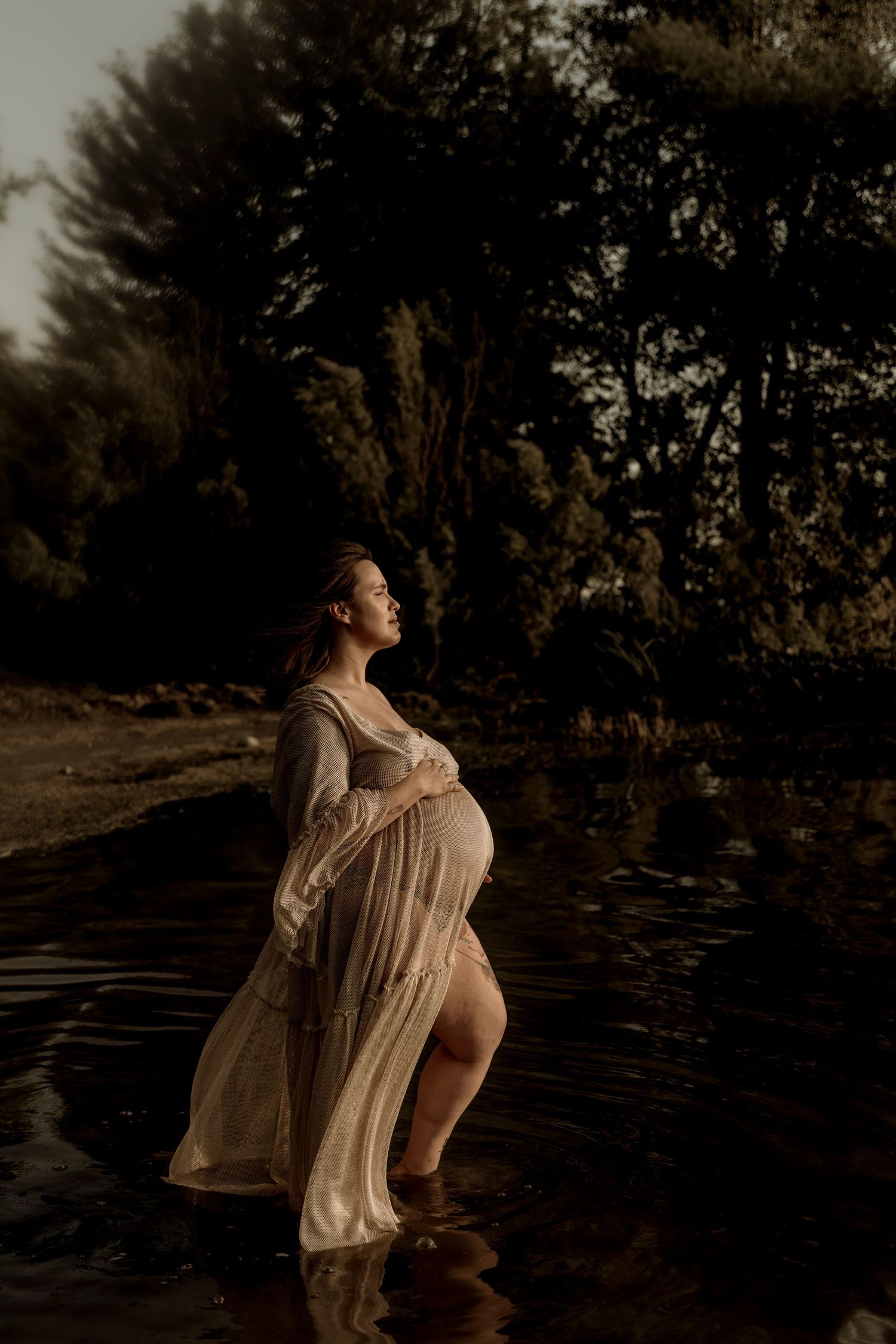 A pregnant woman in a flowing cream dress stands in shallow water at dusk, backlit by golden sunset light through trees.