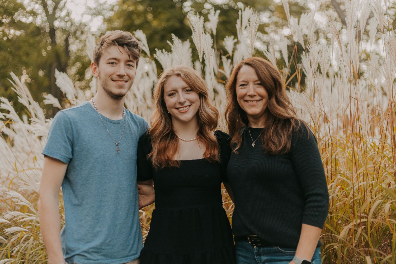 Three people pose together outdoors with tall pampas grass in the background.