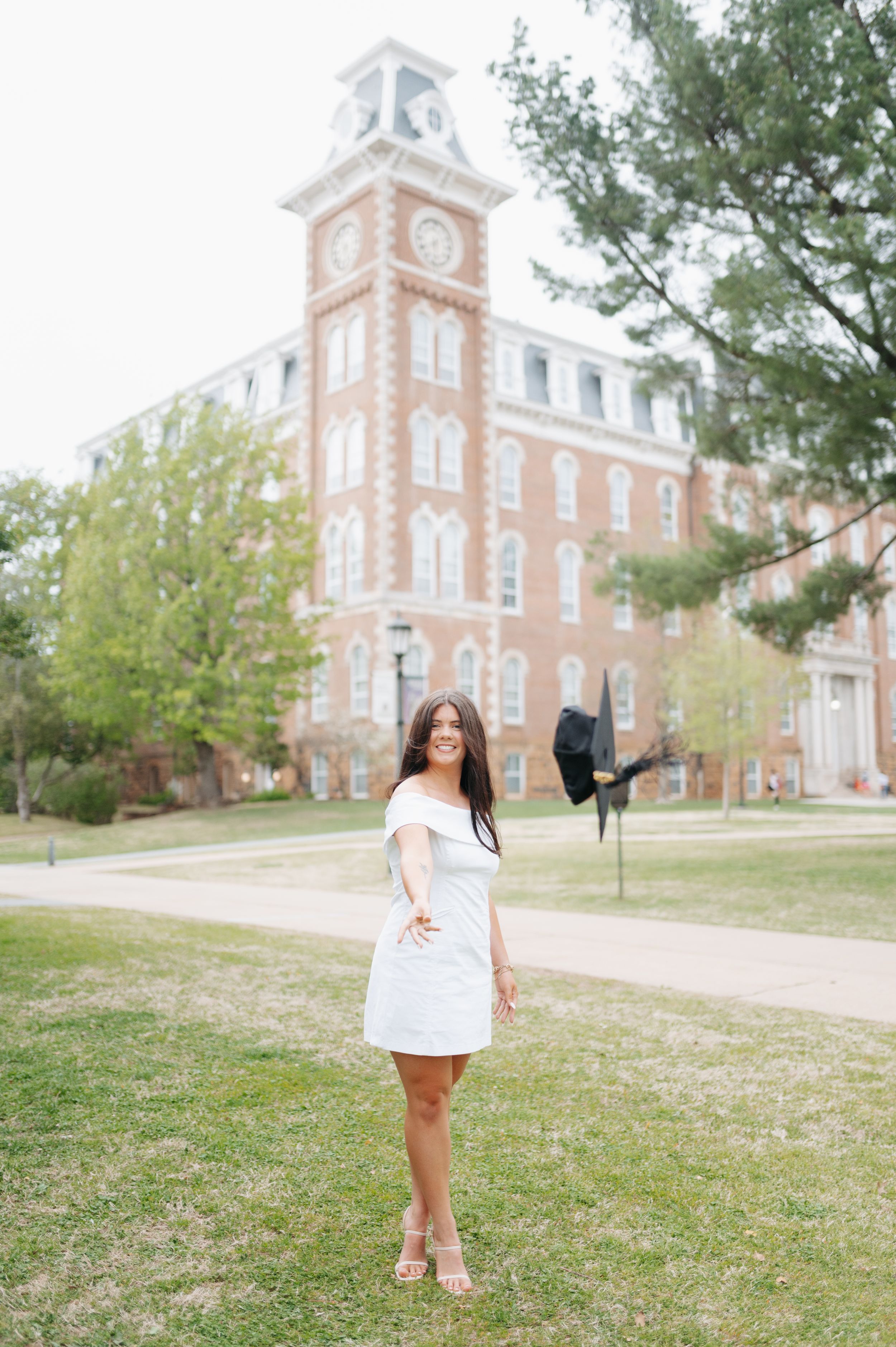 College graduate in white dress tossing cap in front of historic red brick university clock tower.