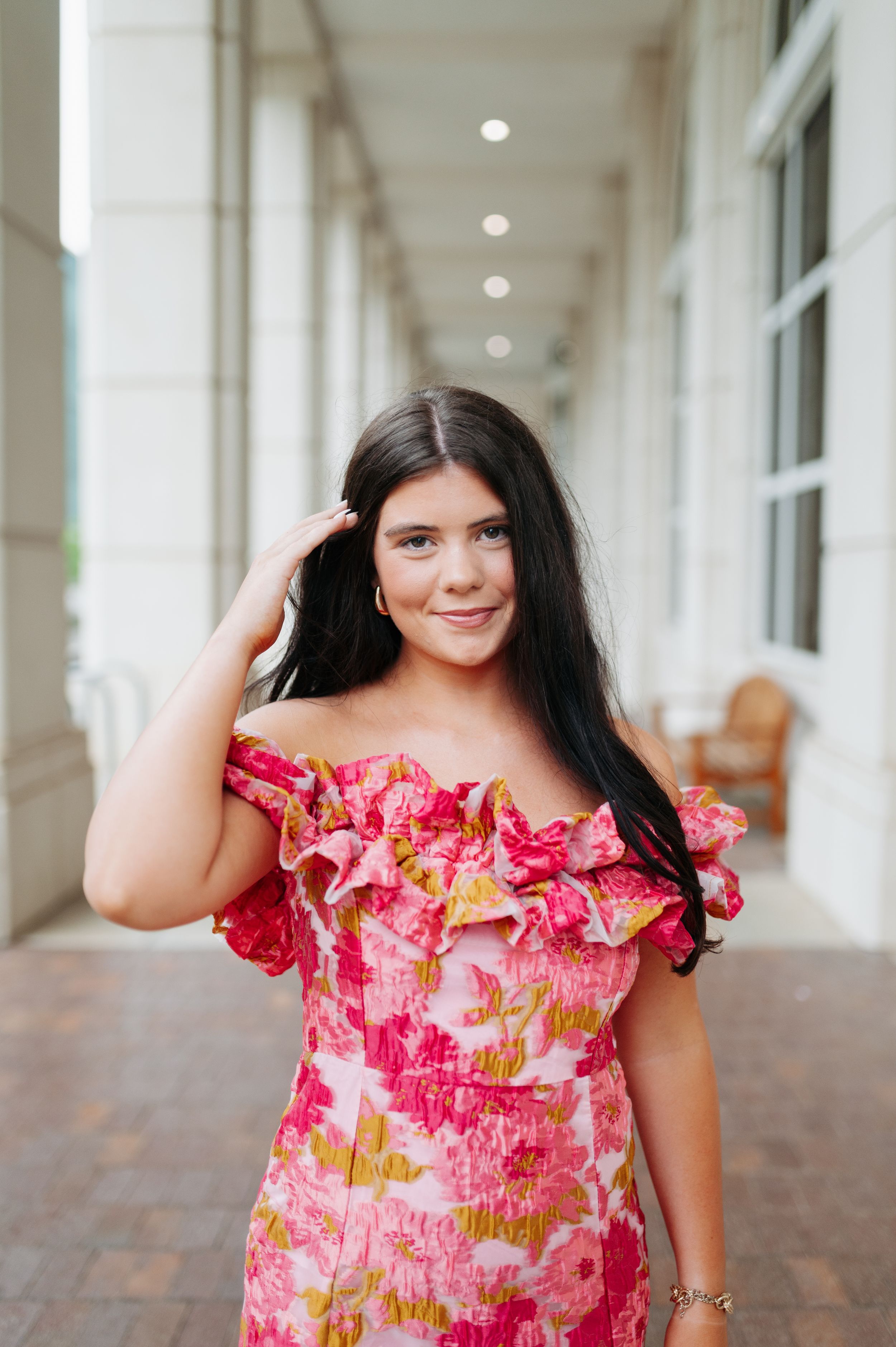 Young woman in pink floral off-shoulder ruffled dress posing in an outdoor corridor, smiling.