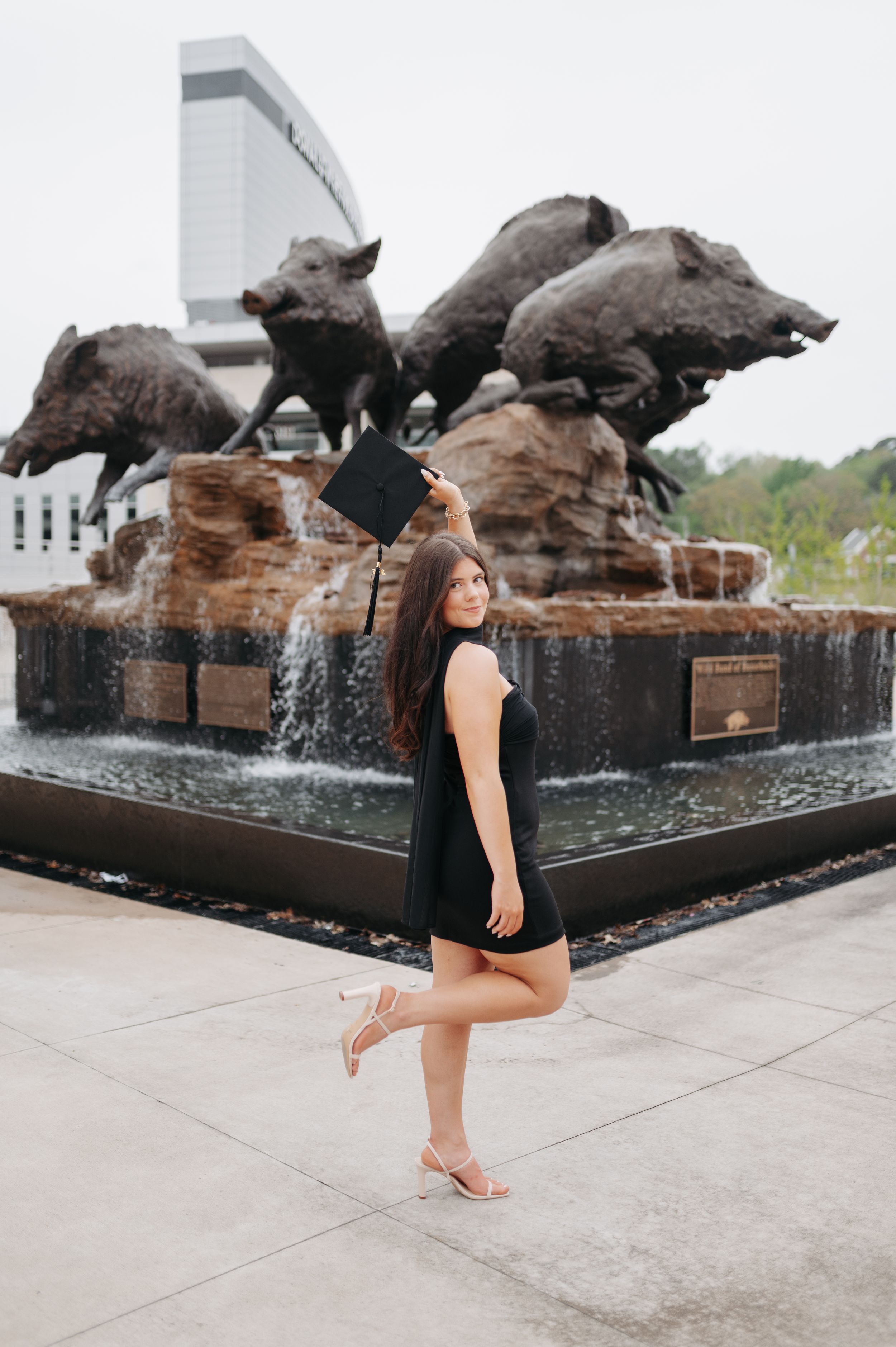 Graduate in black mini dress holding cap beside bronze wild boar fountain sculpture at university.