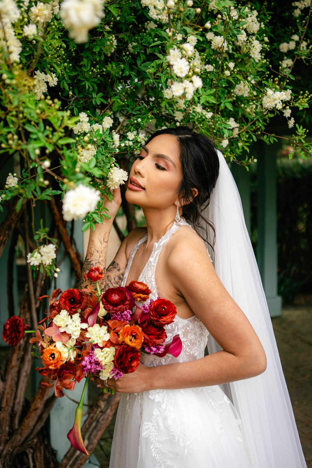 Bride in white gown holds vibrant bouquet against backdrop of blooming white roses.