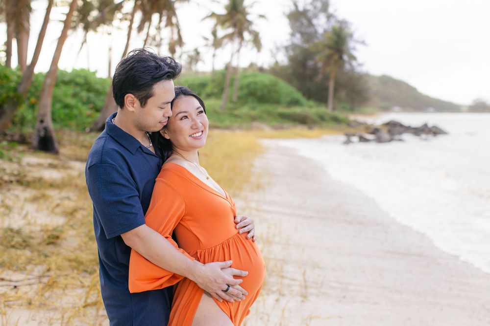 Couple sharing a tender maternity beach moment, woman in orange dress cradling baby bump, palm trees in background.