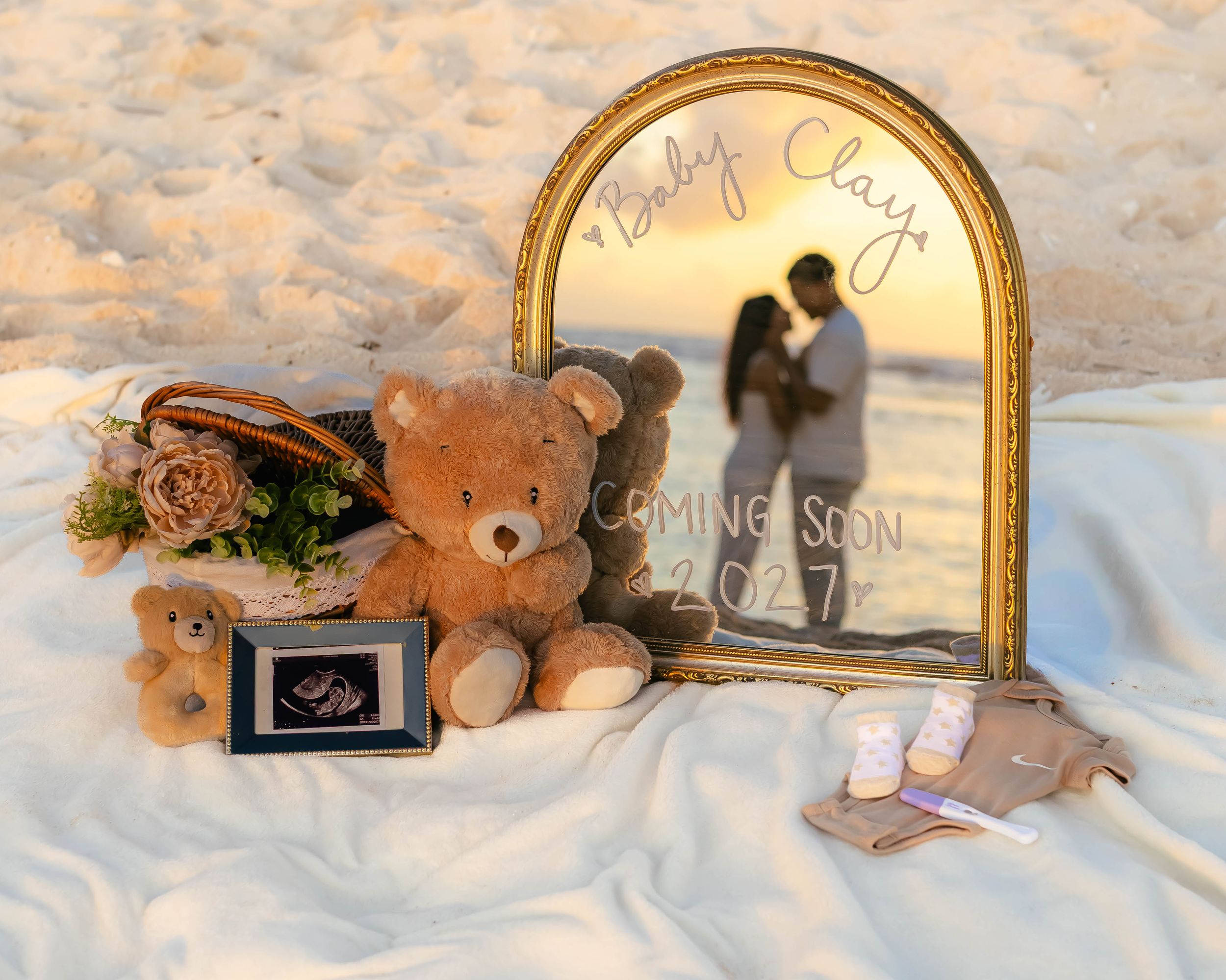 Beach pregnancy announcement with teddy bears, ultrasound photo, and gold mirror reading Baby Clay Coming Soon on white fabric.