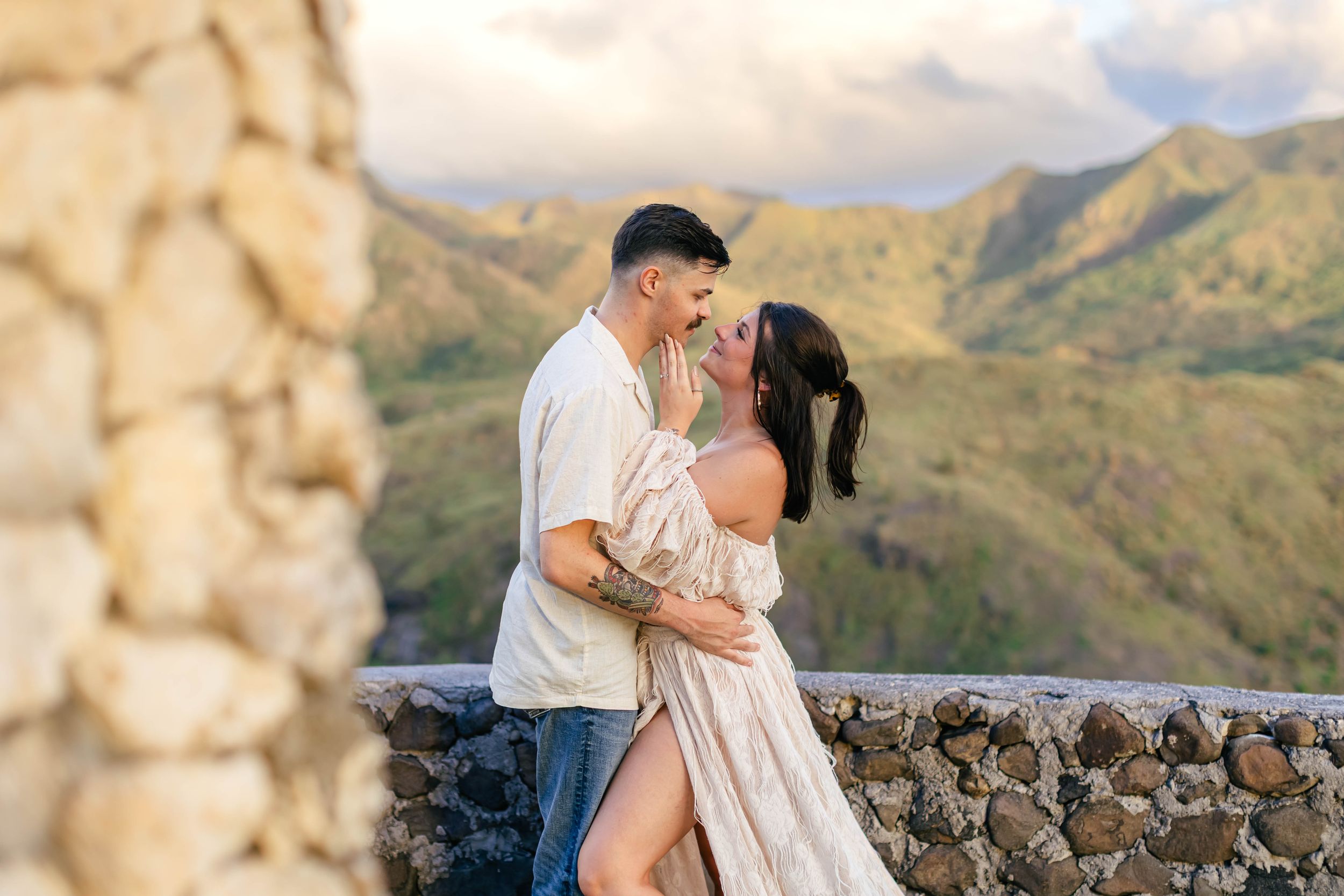 Couple sharing a romantic moment on a stone overlook with lush green Hawaiian mountains in the background.