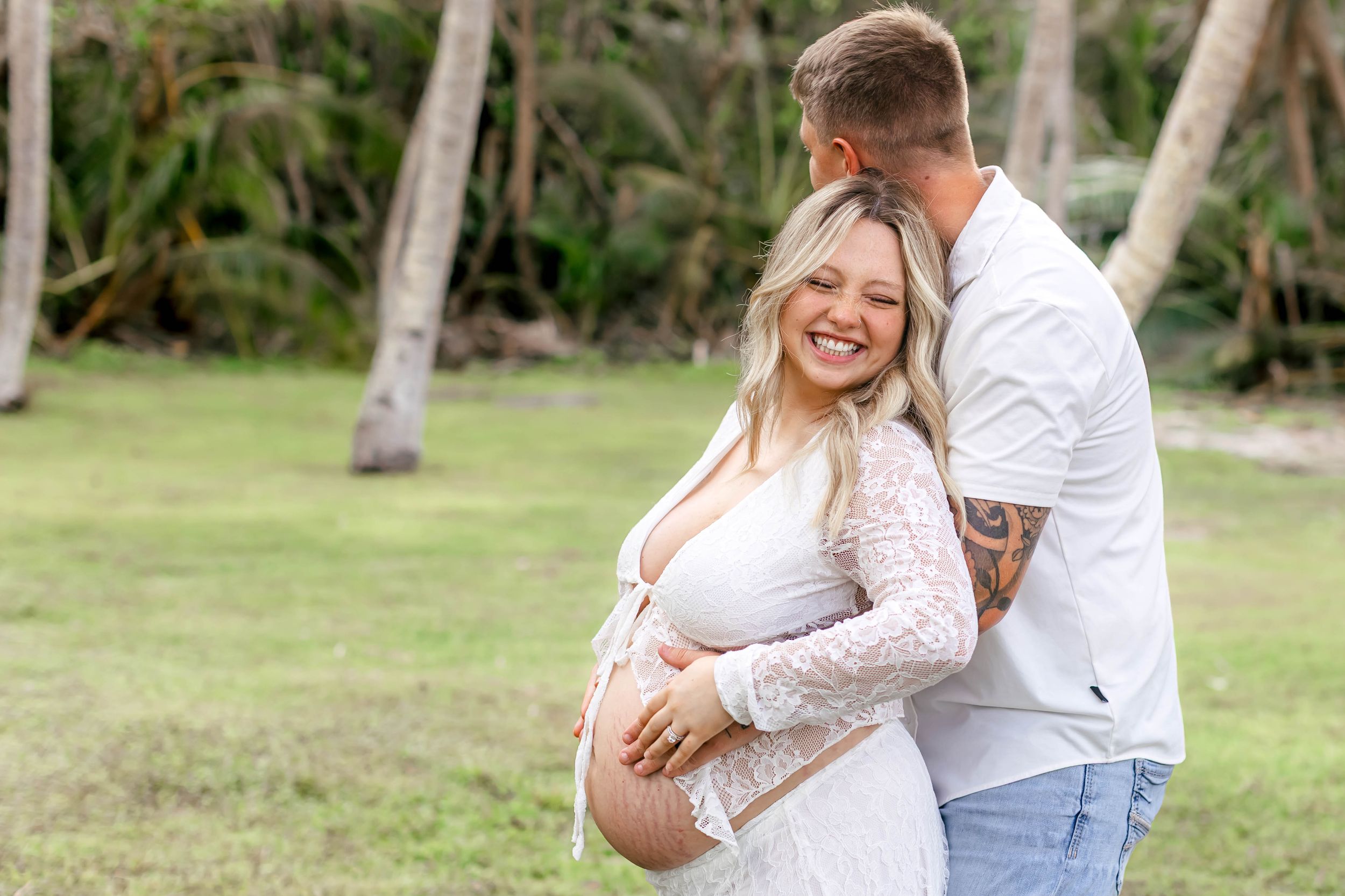 Smiling pregnant woman in white lace dress embraced by tattooed man outdoors near tropical palm trees.