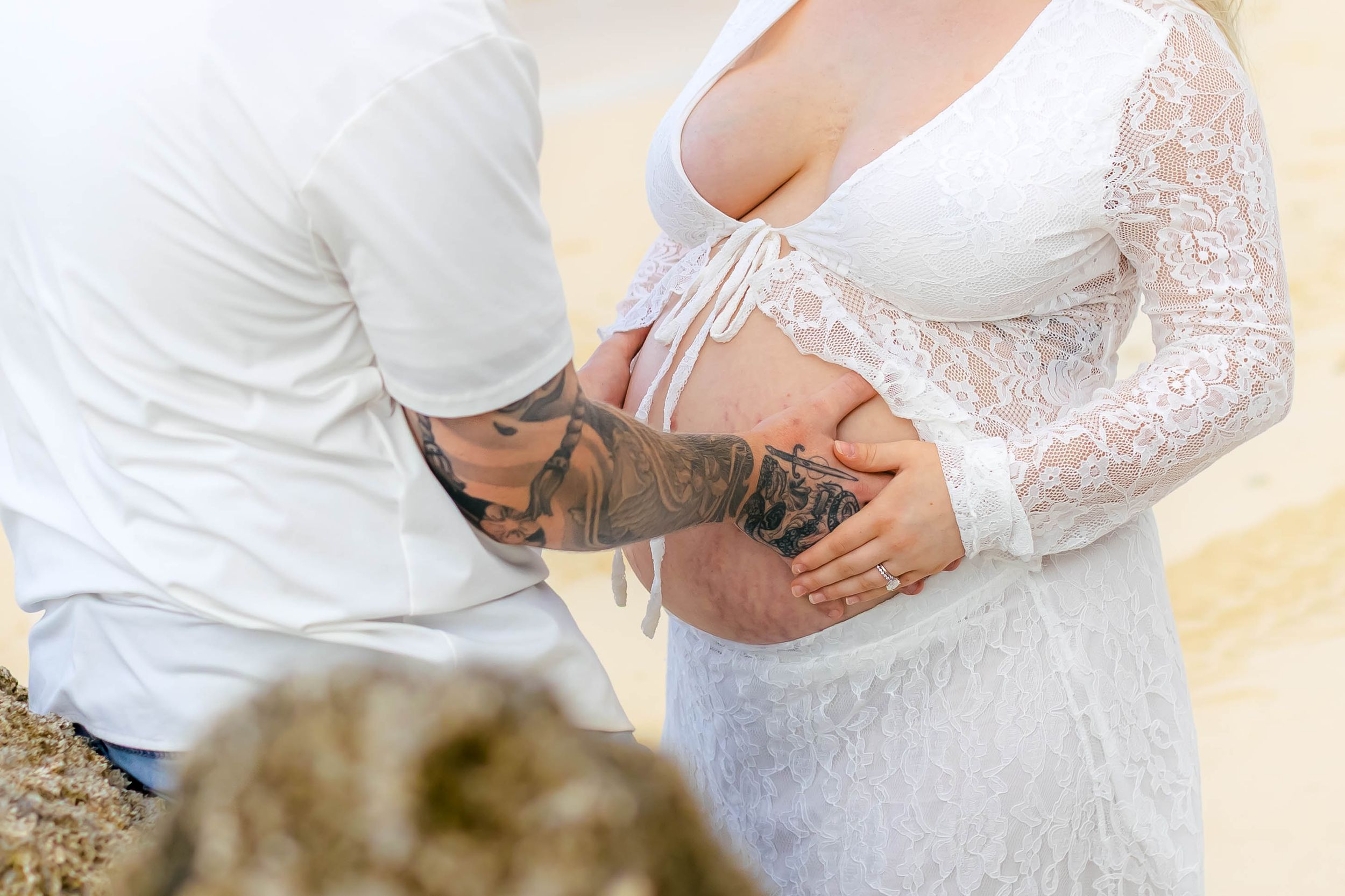 Tattooed man kneeling to kiss pregnant woman's belly in white lace maternity dress during beach photoshoot.