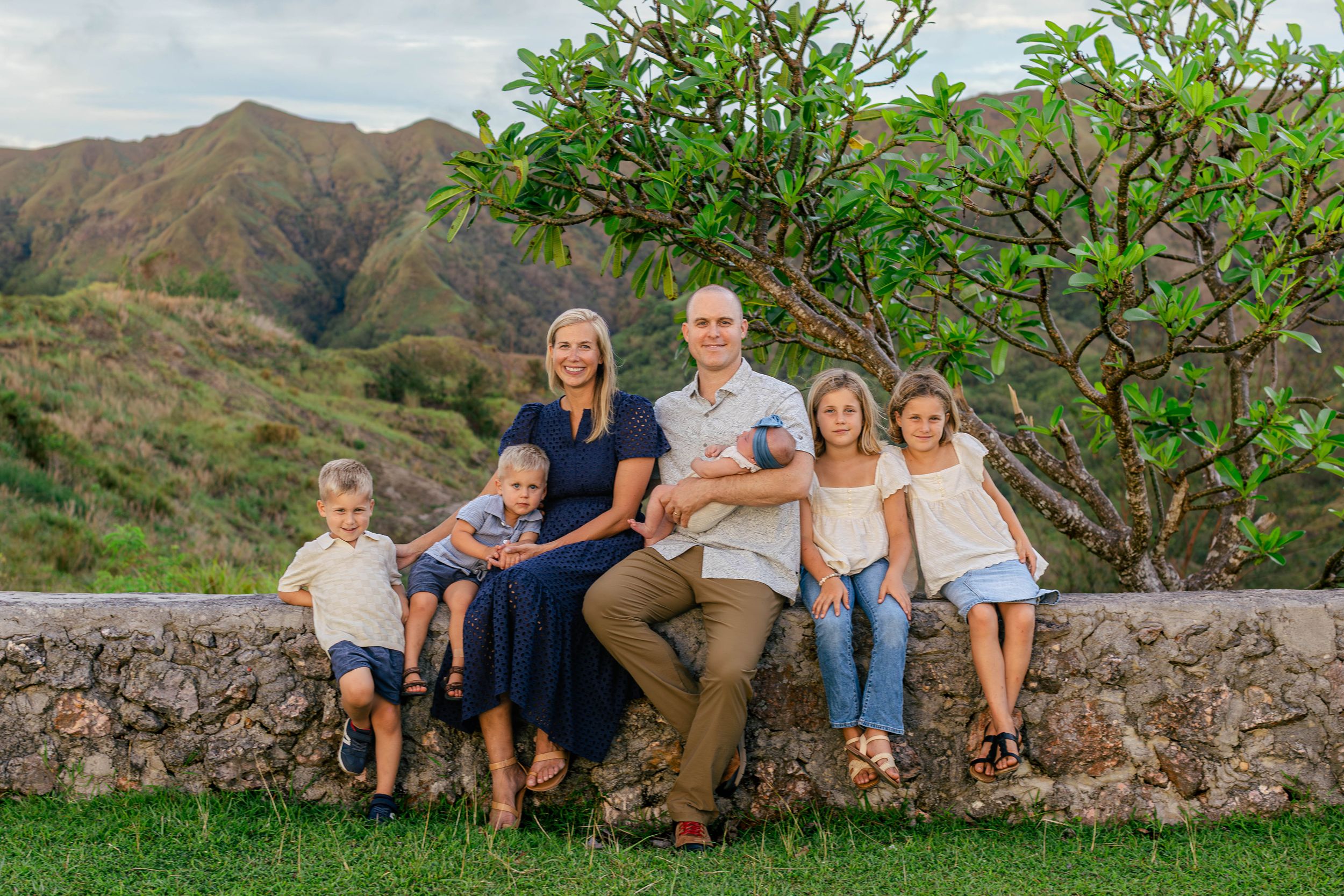 Family of six posing on stone wall with lush green mountains and plumeria tree backdrop in Hawaii.