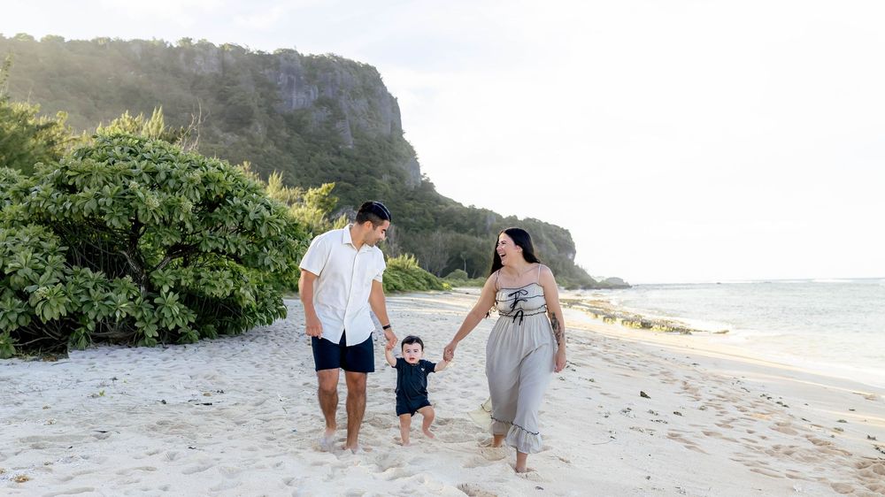 Family enjoying a scenic walk along a tropical beach with mountains and lush greenery in the background.