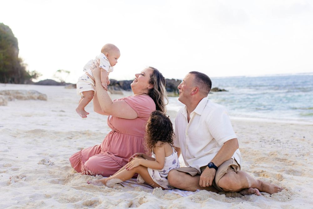Family sitting on sandy beach, parents joyfully lifting baby while young child cuddles nearby with ocean waves in background.