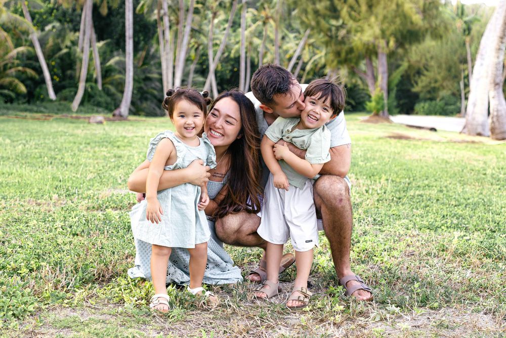 A happy mother crouches with three young children hugging and kissing her in a tropical park setting.