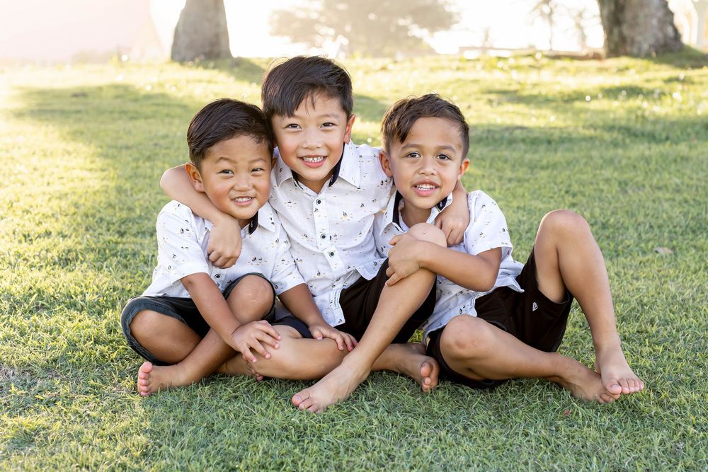 Three smiling boys in matching white shirts and dark shorts sitting together on green grass outdoors.
