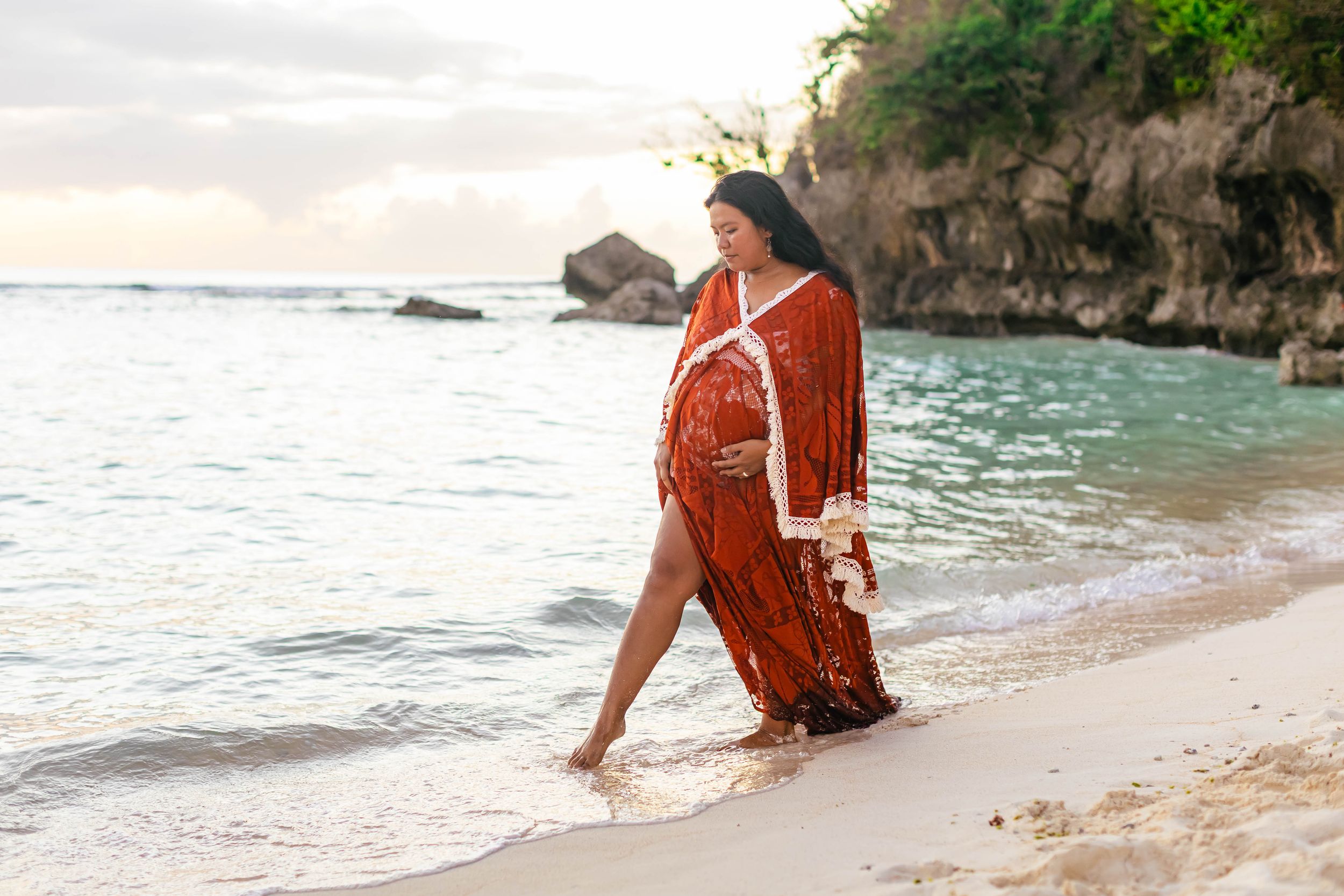 Pregnant woman in rust boho maternity gown walking barefoot on tropical beach at sunset.