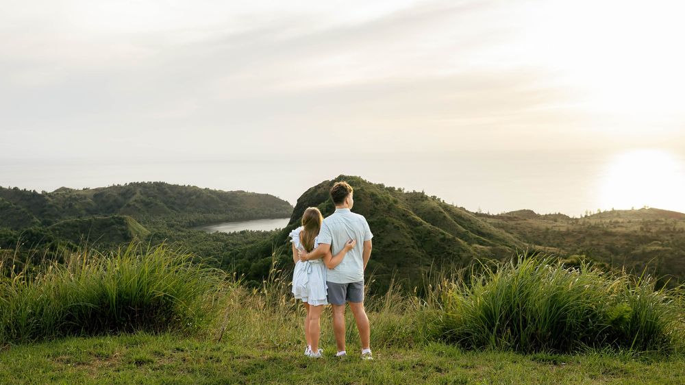 Couple standing together on a lush green hilltop overlooking a scenic coastal valley at sunset.