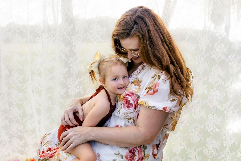 A mother in a floral dress lovingly holds her toddler daughter with a bow, sharing a tender moment indoors.