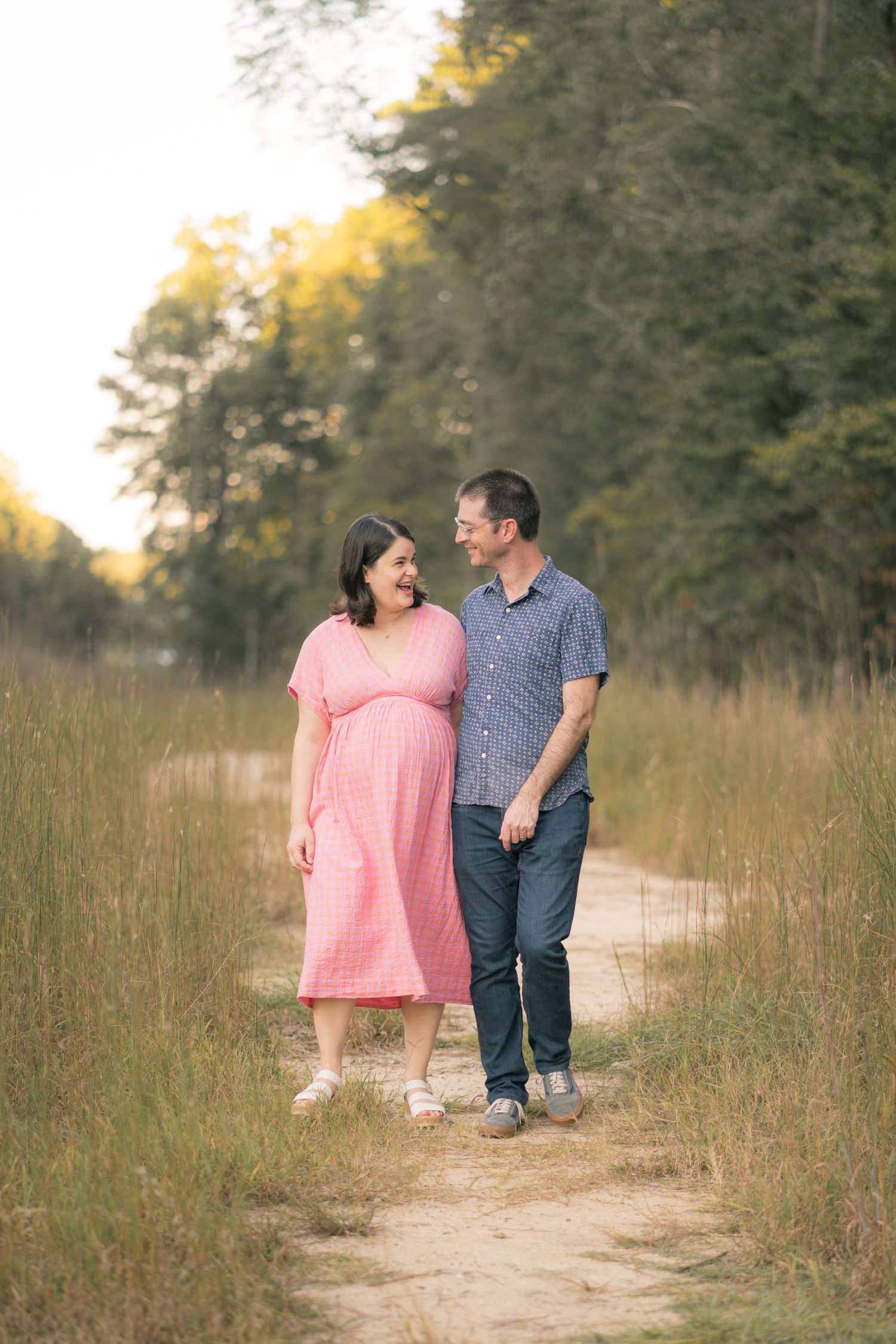 A couple walks together on a dirt path through tall grass during golden hour.
