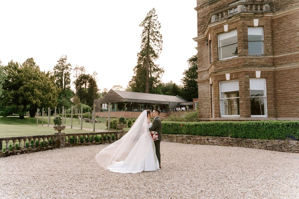 A couple walks on the gravel pathway outside a grand stone manor house on their wedding day.